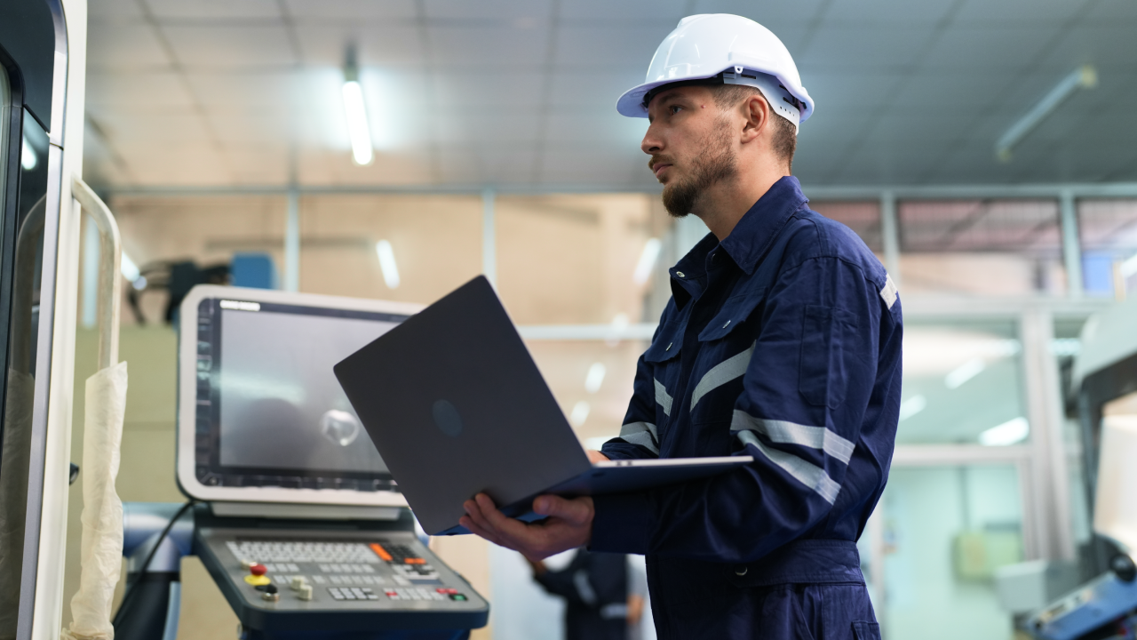 Male engineer using laptop computer operating CNC machine at the factory. Man technician in uniform and helmet safety working at workshop heavy metal industrial