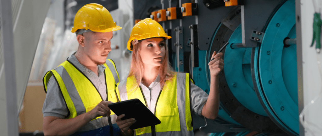 Two factory workers wearing hard hats examining shop floor machines together.