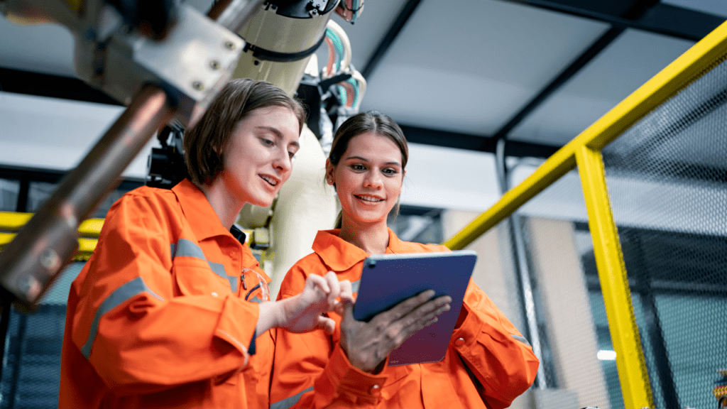 Two female workers in orange overalls, attentively studying data on a tablet, while standing in front of an operational robotic arm in a manufacturing facility.